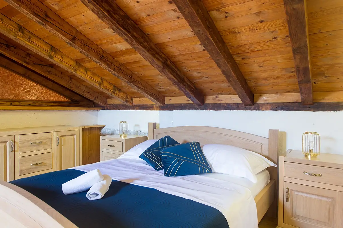 Attic room with a wooden ceiling, a double bed with beautiful bedding and pillows, nightstands on both sides of the bed, a large wardrobe, and lamps on the nightstands.